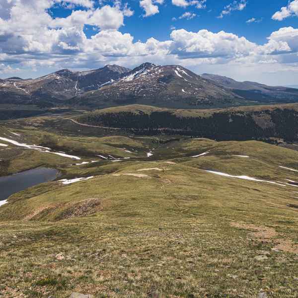 A small dirt path winds down a mountainside into a rolling green valley, an oval Alpine lake to the left, and directly ahead across the valley numerous rocky mountain peaks can be seen. Everything is covered in scattered small patches of snow.