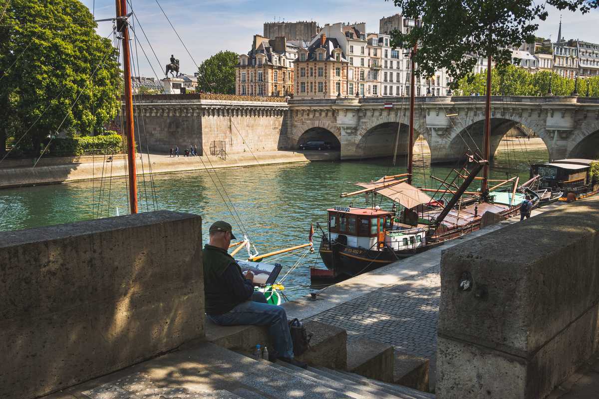 A man sits calmly next to the River Seine painting the scene in the background, an arched stone bridge over the river with several boats docked along the river's edge