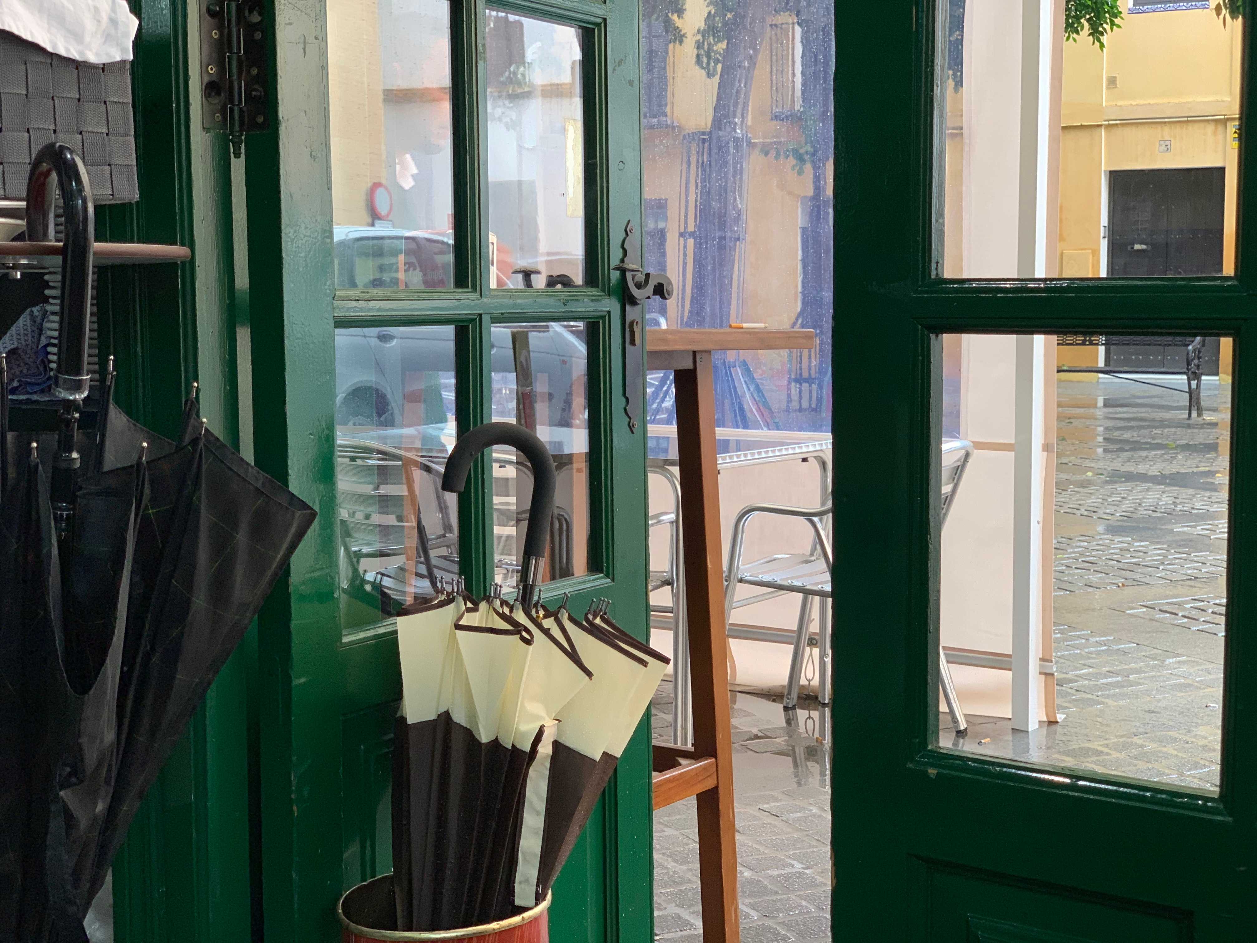 Looking out a green-framed door propped open by an umbrella holder filled with one umbrella on a rainy day. The view outside shows the cafe's outdoor seating on rainy cobblestones